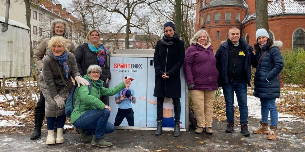 Mehrere Personen stehen neben einer Box auf der Sportbox steht und Menschen mit einem Ball abgebildet sind.