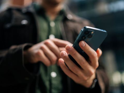 Nahaufnahme der Hände eines nicht identifizierbaren Mannes, der auf einer belebten Straße steht, ein Smartphone hält und bedient – ​​im Internet surft, soziale Medien prüft und eine mobile App nutzt.