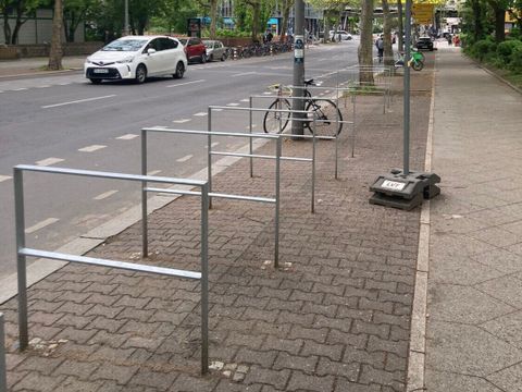 Mehrere silberne Radbügel auf einem gepflasterten Gehweg neben einer Straße mit parkenden Autos und Bäumen