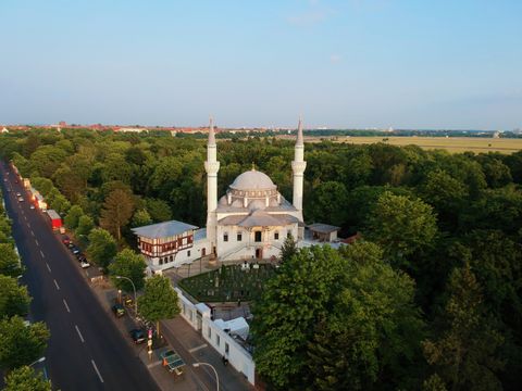 Luftbildaufnahme der Moschee. Weißes Moschegebäude mit einer Kuppel in der Mitte. Rechts und links davon jeweils ein hoher, schlanker Turm. Die Moschee ist umgeben von vielen dunkelgrünen Bäumen
