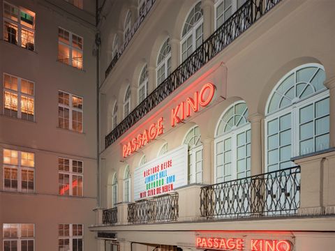 Es ist die Neuon-Leuchtreklame der Passage Kinos zu sehen. Die Leuchtreklame ist auf der Hauswand angebracht. In roten Neon-Buchstaben leuchtet der Schriftzug "Passage Kino". Darunter auf einem weißen Steckboard sind in roten, blaune und grünen Schriftzeichen die aktuellen Filmtitel gesteckt, die das Kino zeigt.