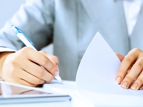 Eine Person in formeller Kleidung hält ein Blatt Papier und schreibt mit einem Stift darauf, vermutlich beim Ausfüllen oder Unterschreiben eines Dokuments.