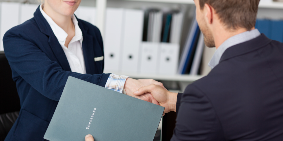 Eine Frau und ein Mann begrüßen sich per Handschlag in Businessgarderobe. Der Mann hält eine Bewerbungsmappe in der Hand.