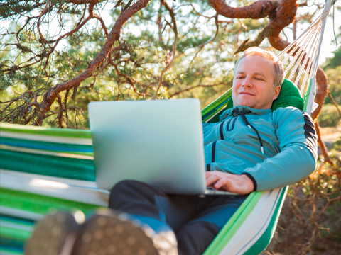 Ein Mann liegt in einer Hängematte im Wald und guckt auf einem Laptop
