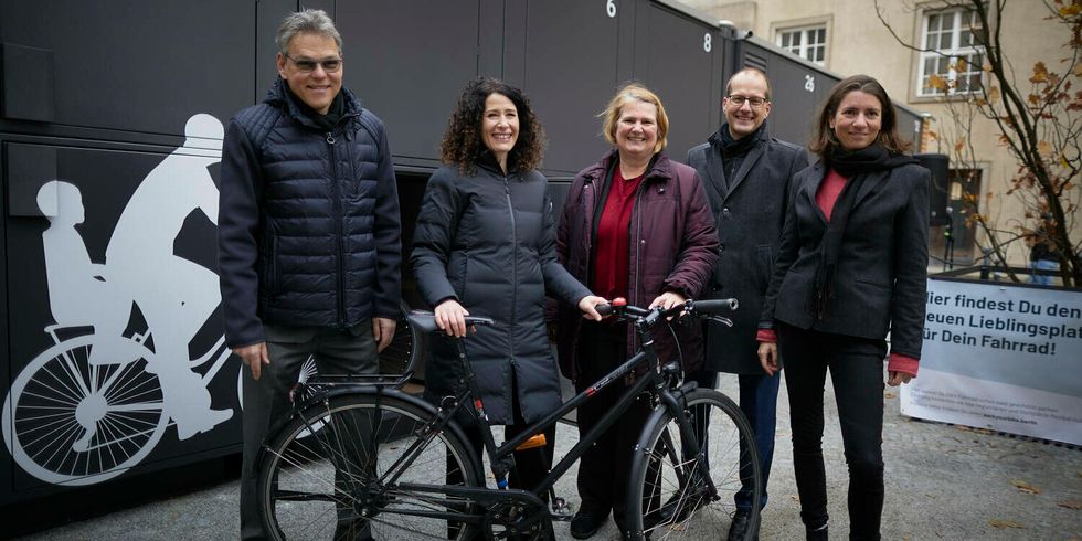 Fünf Personen stehen zusammen mit einem Fahrrad vor einem Container mit nummerierten Abstellplätzen.