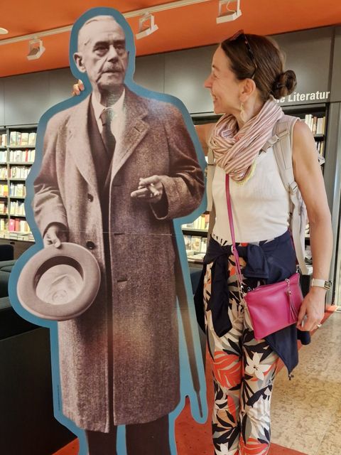Iris Schaub lächelt eine Pappfigur von Thomas Mann an. Sie trägt dabei eine bunte Hose.