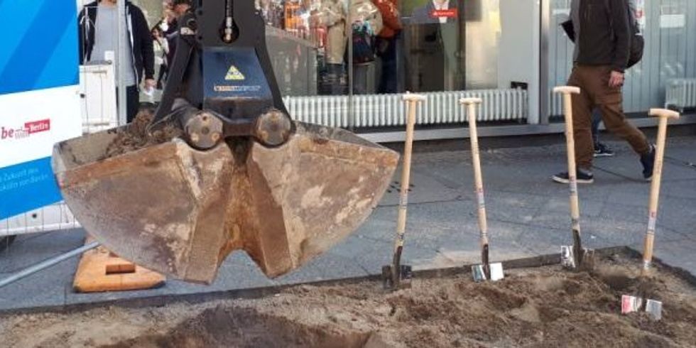 Foto: neben einer Baggerschaufel stecken vier Spaten im sandigen Boden, im Hintergrund ein Gehweg und Schaufenster