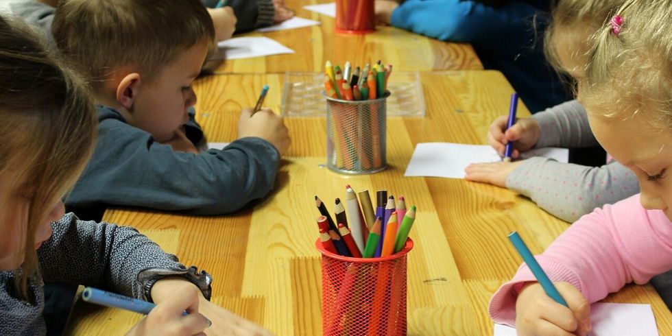 Kinder sitzen an einer Tischreihe und malen mit Buntstiften.