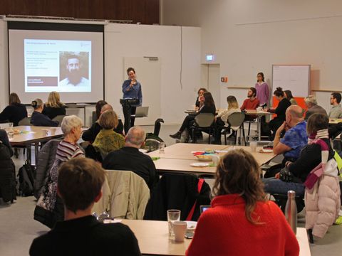 mehrere Personen sitzen an Tischen und schauen sich auf einem Bildschirm einen Vortrag an