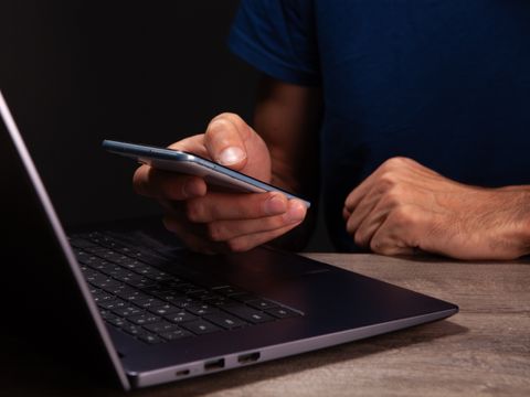 Laptop im Vordergrund, zwei Hände zu sehen mit Handy in der rechten Hand