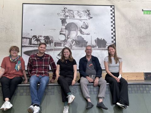 Fünf Personen sitzen nebeneinander auf dem Rand einer Skateboard-Rampe in einer Halle. Im Hintergrund hängt ein großes Schwarz-Weiß-Foto, das einen Skateboarder bei einem Trick zeigt. Die Gruppe wirkt entspannt und lächelt in die Kamera. Die Wände sind schlicht, und über dem Foto ist ein Notausgangsschild zu sehen.