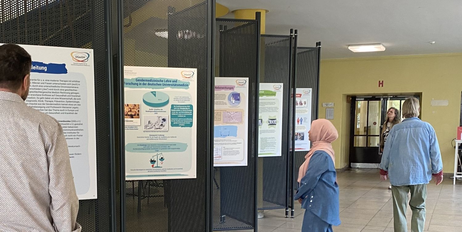 Vor den Ausstellungswänden der Ausstellung über gendersensible Medizin der VHS Neukölln stehen drei Personen an verschiedenen Stellen und schauen sich die Poster an den Stellwänden an.