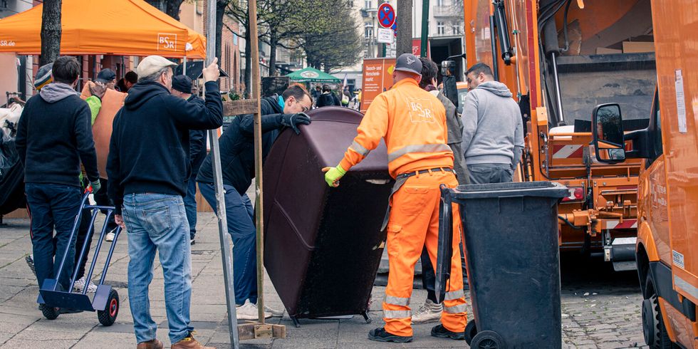 Auf einem Gehweg tragen zwei Männer eine Couch. Einer der Männer trägt die Uniform der BSR. Am Straßenrand sind BSR-Wägen abgestellt und im Hintergrund sind weiter Menschen zu erkennen, einer zieht einen Handwagen hinter sich her.