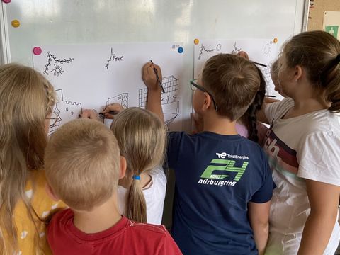 Sieben Kinder zeichnen zusammen an zwei großformatigen Malvorlagen, die an der Wand hängen.