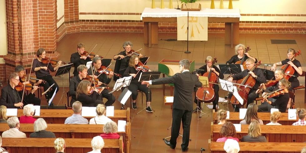 Ein Orchester führt in einer Kirche ein Stück auf. Auf den Kirchenbänken sitzen Menschen und schauen und hören zu.
