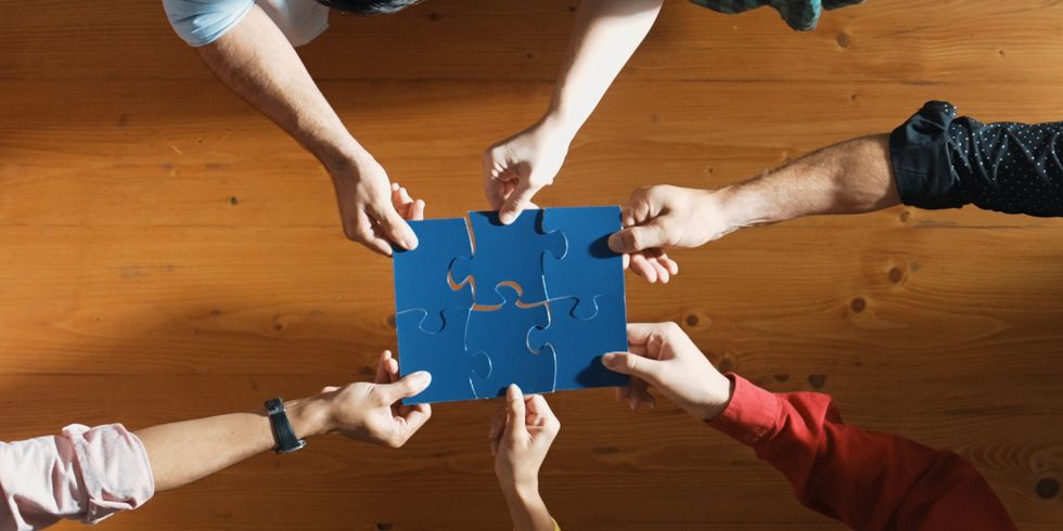 Sechs Unterarme sind in der Draufsicht zu sehen. Die zugehörigen Hände halten jeweils ein blaues Puzzleteil. Sie fügen die Puzzleteile in der Mitte so zusammen, dass sie ineinanderpassen.
