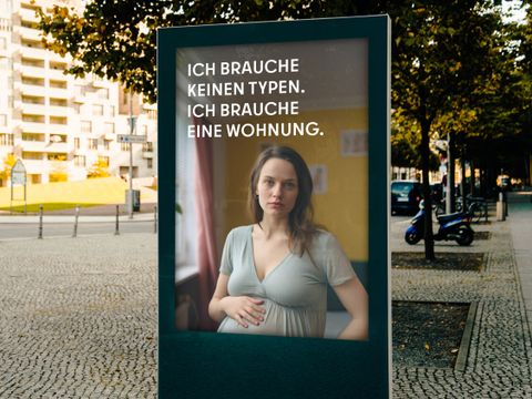 Auf einer Stadtwerbetafel ist eine schwangere Frau abgebildet mit dem Text: “ICH BRAUCHE KEINEN TYPEN. ICH BRAUCHE EINE WOHNUNG.” Im Hintergrund sind Bäume, Gebäude und ein Gehweg zu sehen.