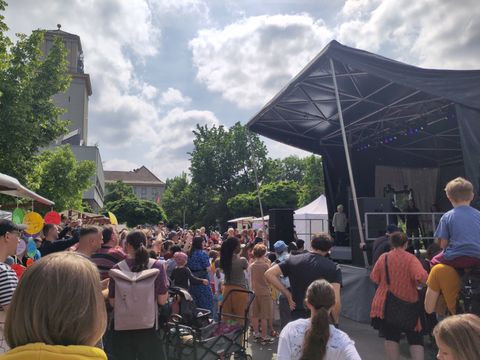 viele Menschen stehen vor einer Bühne und schauen auf diese. Im Hintergrund Marktstände und das Rathaus Tempelhof, Bäume und Wolken