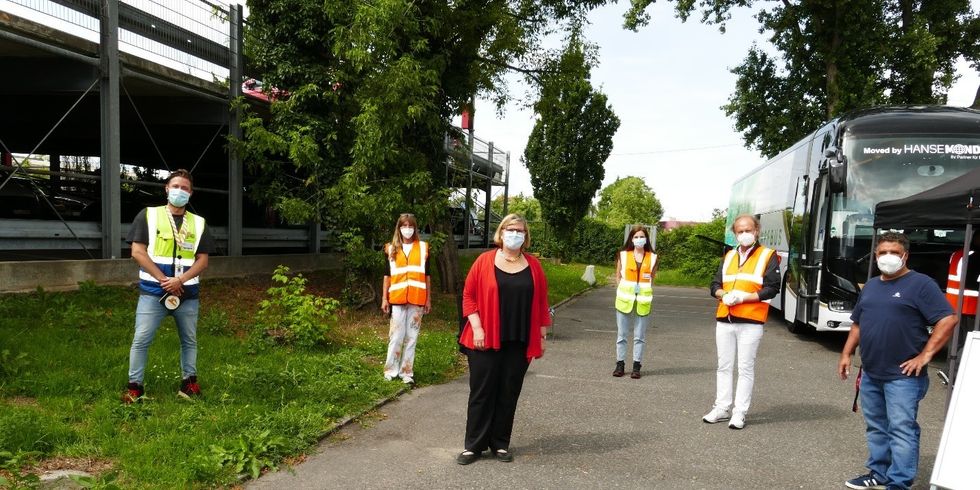 Sechs Personen stehen mit Masken und Abstand zueinander auf einem Parkplatz. Hinter Ihnen ist ein Bus, der sogenannte Impftruck.