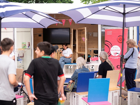 Ein Vortrag wird im Freien gehalten, das Publikum sitzt und steht unter Sonnenschirmen