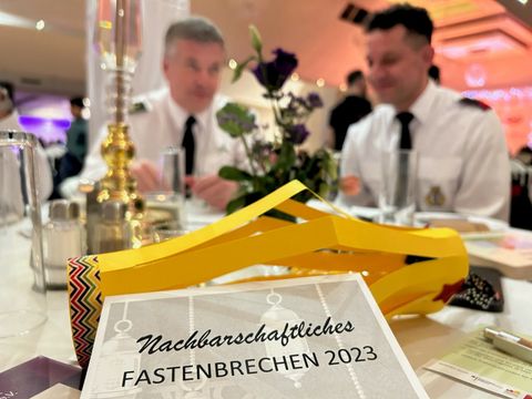 Geschmückter Tisch, im Vordergrund ein Flyer für das Nachbarschaftliche Fastenbrechen 2023, im Hintergrund zwei Gäste der Veranstaltung