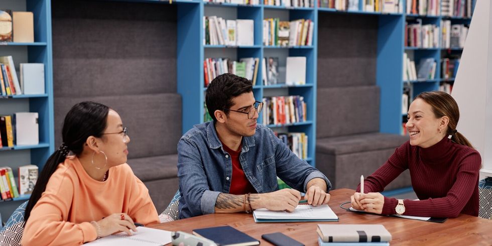 Drei Studierende sprechen miteinander in der Bibliothek an einem Tisch.