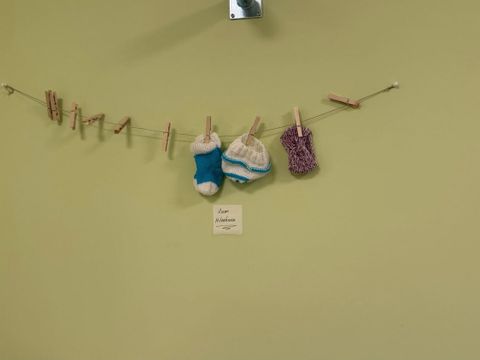 An einer hellgrünen Wand im FamilienServiceBüro Friedrichshain-Kreuzberg ist eine Leine mit Holzklammern befestigt. Daran hängen selbstgestrickte Kleidungsstücke in verschiedenen Farben, darunter Mützen und Söckchen. Unter der Leine befindet sich ein kleiner Zettel mit handschriftlichem Text "Zum mitnehmen".
