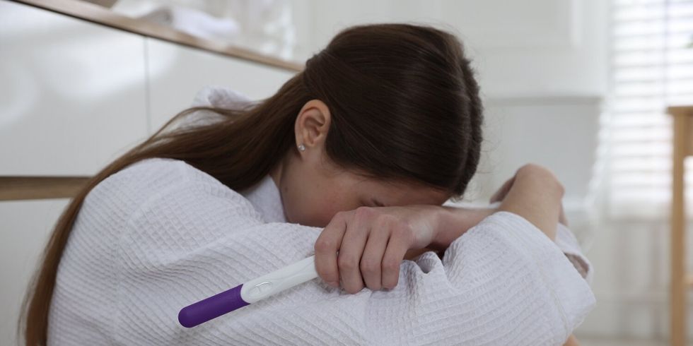 Eine Frau sitzt auf dem Boden, den Kopf auf ihre verschränkten Arme gelegt, wirkt traurig oder besorgt und hält einen Schwangerschaftstest in der Hand.