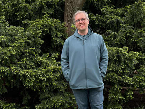 Aaron Schaar steht mit leicht nach vorne geneigtem Oberkörper vor dichtem, sattgrünem Nadelgehölz. Er trägt eine hellblaue Jacke mit Reißverschluss und eine dunkelblaue Jeans. Die Hände sind in den Jackentaschen. Der Hintergrund wird von den feinen, frischen Trieben der Nadelbäume dominiert. Ein dicker Baumstamm ist hinter ihm sichtbar.