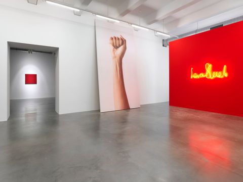In einem weißen Raum mit einem Betonfußboden sieht man auf der rechten Seite eine rote Wand mit einem leuchtenden Schriftzug. Links daneben ein übermenschgroßes Bild eines ausgestreckten Arms mit einer geballten Faust