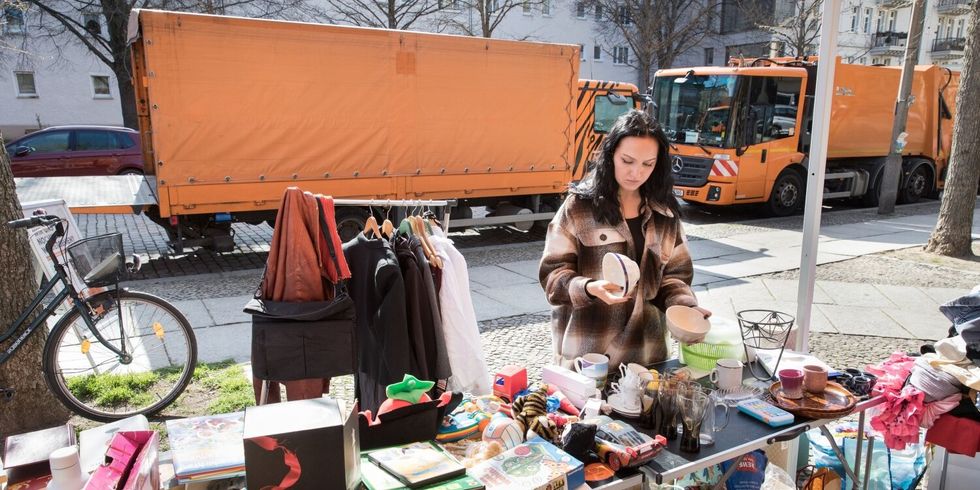 Eine Frau steht an einem Straßenrand an einem Stand mit unterschiedlichen Dingen. Hinter ihr stehen zwei große Wägen der Berliner Stadtreinigung.