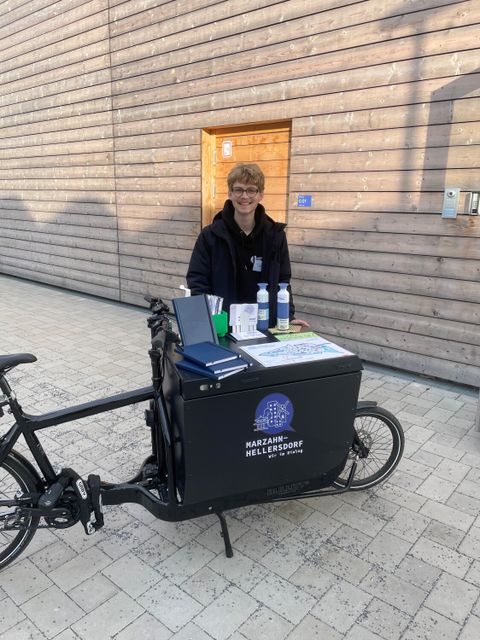 Jonas vom Raum für Beteiligung steht mit dem Beteiligungsrad vorm Eingang zur Einwohnerversammlung. Auf dem Fahrrad befinden sich Goodies und die Karte der Bezirksregion.