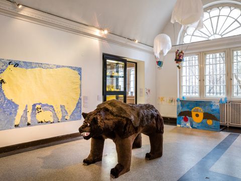 Im Vordergrund eine Bärenfigur in Lebensgröße. Oberhalb der Bärenfigur zwei Ballons über die ein weißes Tuch gespannt ist und ein Korb unten hängt. Es soll aussehen wie ein Heißluftballon. inter der Bärenfigur an der Wand eine große Zeichnung, die ebenfalls ein Tier zeigt. Es ist in gelb gezeichnet auf blauem Hintergund. Es handelt sich um Kunstinstallationen, die von Kindern gemacht wurden .