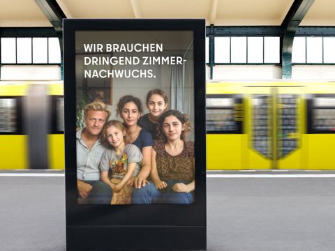 Das Bild zeigt eine Werbetafel auf einem Bahnsteig. Die Werbetafel zeigt eine Familie, die aus einem Mann, einer Frau und drei jungen Mädchen besteht. Auf der Tafel steht: “WIR BRAUCHEN DRINGEND ZIMMERNACHWUCHS.”
