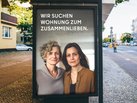 Das Bild zeigt eine Werbetafel an einer Straße. Auf der Tafel sind zwei Frauen abgebildet, die nebeneinander stehen und in die Kamera blicken. Der Text auf der Tafel lautet: “WIR SUCHEN WOHNUNG ZUM ZUSAMMENLIEBEN.” Im Hintergrund sind Gebäude, eine Straße und ein Fußgänger zu erkennen.