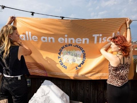 Zwei Frauen halten ein Banner mit der Aufschrift alle an einen Tisch hoch