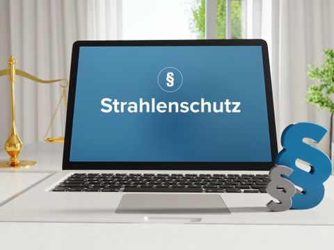 Symbolgrafik mit dem Begriff Strahlenschutz auf einem Laptopmonitor mit Paragraphen und Balkenwaage als Symbole daneben auf einem Tisch