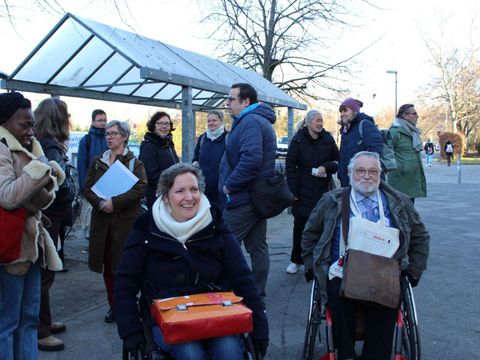 Menschen stehen in mehreren Grüppchen zusammen und unterhalten sich angeregt. Im Vordergrund sind ein Mann und Frau im Rollstuhl und schauen lachend in die Kamera.