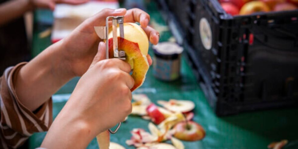Hände die einen Apfel schälen, im Hintergrund eine Kiste Äpfel und bereits viele Schalenreste auf dem Tisch