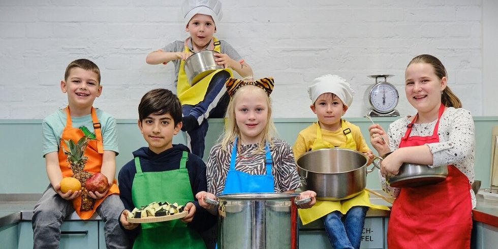 Kinder sitzen und stehen in einer Küche. Sie tragen Schürzen und halten Töpfe, Teller und Obst in der Hand.