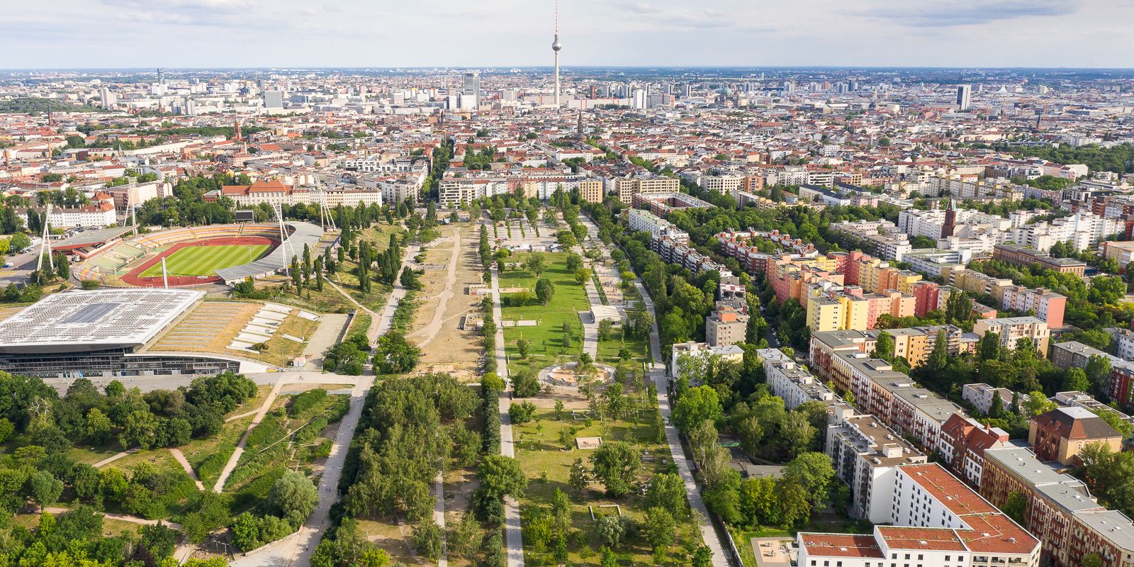 Das Bild zeigt eine Luftaufnahme vom Berliner Mauerpark. Im Vordergrund ist eine große, grüne Parkanlage mit Wegen, Bäumen und Wiesen zu sehen. Links befinden sich ein Stadion und weitere Sportanlagen. Im Hintergrund erstreckt sich die dichte Stadtbebauung, mittig ragt der Berliner Fernsehturm empor.