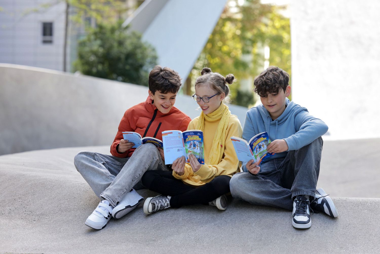 Fotografie von drei lesenden Kindern, die mit Büchern draußen auf dem Boden sitzen