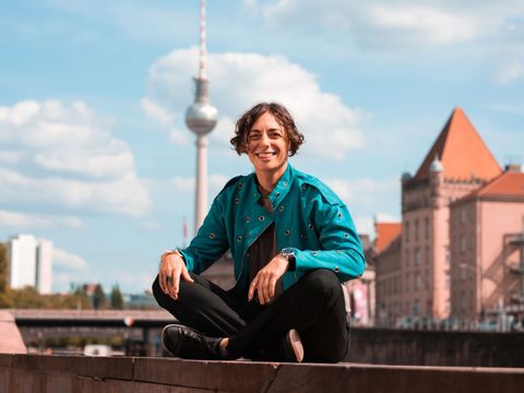 Alex M. Gastel sitzt lachend auf einer Mauer. Im Hintergrund ist der Berliner Fernsehturm zu sehen.