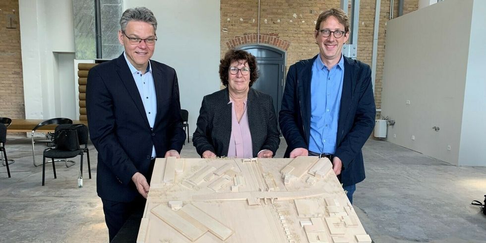 Drei Personen stehen zusammen hinter einem Tisch und lächeln in die Kamera. Zusammen heben sie eine Holzplatte an auf der ein Teil einer Stadt aus der Vogelperspektibe eingeschnitzt ist.