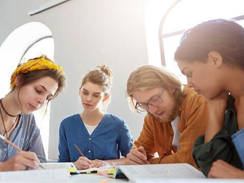 Mehrere Personen sitzen mit Stiften in den Händen an einem Tisch und lesen in Heften, die auf dem Tisch liegen