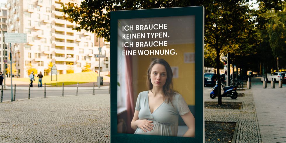 Auf einer Stadtwerbetafel ist eine schwangere Frau abgebildet mit dem Text: “ICH BRAUCHE KEINEN TYPEN. ICH BRAUCHE EINE WOHNUNG.” Im Hintergrund sind Bäume, Gebäude und ein Gehweg zu sehen.
