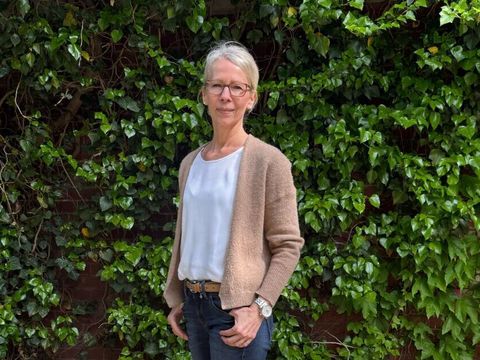Anja Guth steht im Freien vor einer dichten, grünen Wand aus Efeu. Sie trägt eine beigefarbene Strickjacke über einem weißen Oberteil, eine dunkle, schmal geschnittene Hose und beigefarbene Schuhe. Ihr linker Arm ist locker angewinkelt, die rechte Hand ruht in der Hosentasche. Der Untergrund besteht aus hellem, strukturiertem Boden.