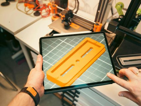 Auf einem Tablet ist ein 3-D Modell zu sehen, im Hintergrund ein 3-D-Drucker