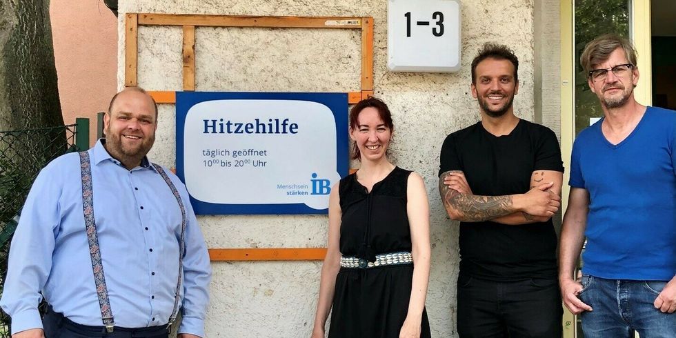 Drei Personen stehen vor einem Gebäüde, an dem ein Schild angebracht ist, auf dem Hitzehilfe steht.