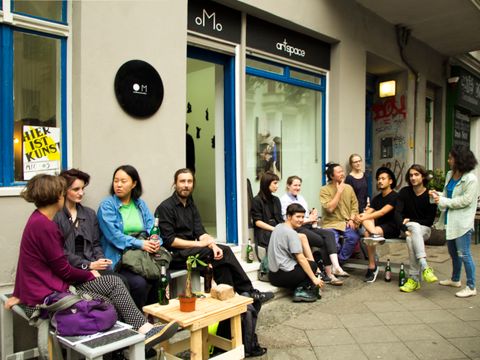Vor einem Ladenlokal sitzen rechts und links von der Tür an niedrigen Tischen und Stühlen mehrere Personen und unterhalten sich
